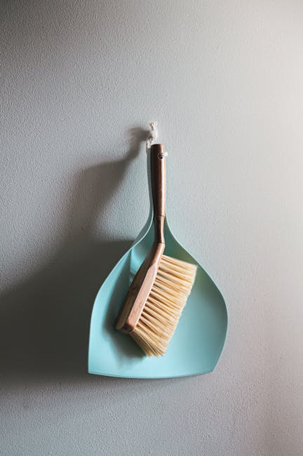 A dustpan and brush hanging on a white wall, with the dustpan in soft teal color and the brush featuring a wooden handle and light-colored bristles. The tools are positioned together, with the brush r