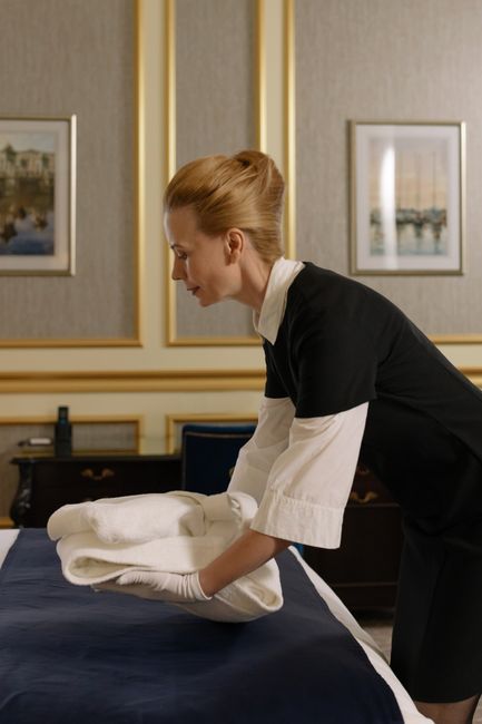 A professional housekeeper with red hair tied back is arranging clean, white linens on a dark blue bed in a well-lit, elegantly decorated bedroom. The room features beige and gold framed wall art, with a wooden dresser and a bedside table visible in the background. She is dressed in a black and white uniform, wearing white gloves, and her focus is on ensuring the bedding is perfectly arranged for a tidy, hygienic appearance. This scene illustrates thorough domestic surface cleaning and bed linen sanitisation as part of comprehensive end of tenancy cleaning services offered by Cleaners W14, emphasizing attention to detail in maintaining cleanliness and hygiene in residential spaces in SW13, W14.