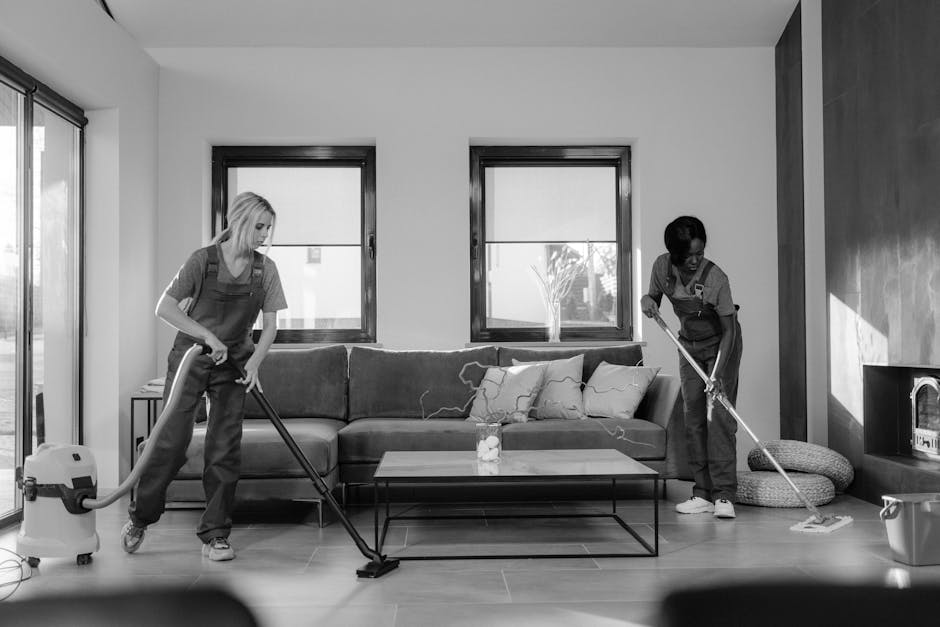Two professional cleaners from Cleaners W14 are performing surface cleaning in a modern living room. They are using vacuum and mop tools to clean the wooden flooring, which appears to be free of dust and debris, showcasing a polished and tidy appearance. The room features a sleek, contemporary style with a dark-toned sofa, decorative cushions, and large windows that let in natural light, illuminating the space. The background includes a coffee table with a small decorative item, a stone fireplace, and some cleaning supplies placed on the floor. The scene reflects thorough domestic cleaning services focused on maintaining hygiene and surface cleanliness in a residential setting, aligning with Barnes Village end of tenancy cleaning services SW13, W14 offered by Cleaners W14.