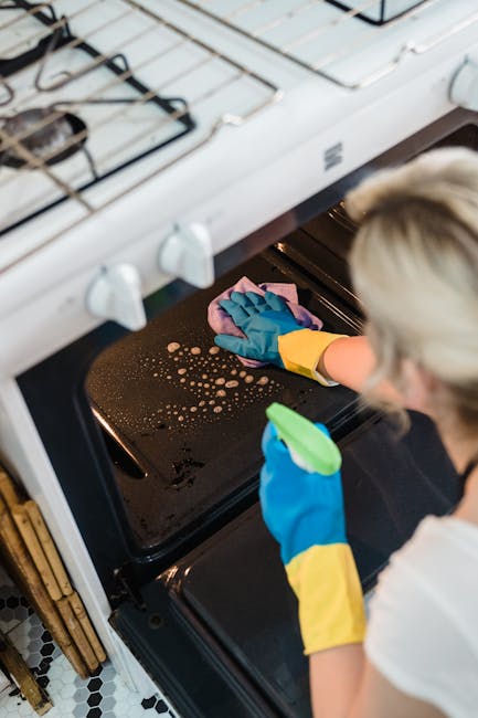 A person wearing yellow and blue rubber gloves is cleaning the black glass surface of an oven with a pink microfiber cloth, removing burnt-on residue and grease stains. The oven door is open, revealing the interior racks. Above, a white gas stove with metal burners and control knobs on a tile countertop is visible. The scene is illuminated by natural light, highlighting the cleanliness and shine of the oven surface, indicating a thorough domestic kitchen deep cleaning service. This image exemplifies surface cleaning and sanitisation performed by Cleaners W14 as part of their kitchen deep clean on Church Road in Barnes, W14.