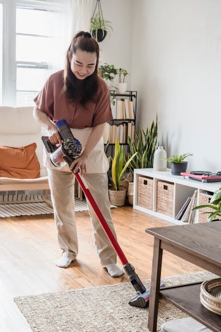 A woman with long dark hair tied back, dressed in a brown top and beige trousers, is engaged in surface cleaning of a wooden floor in a well-lit, modern living room. She is using a handheld vacuum cleaner with a red tube and a black cleaning head to vacuum a beige area rug. The room features a white wall with a large window allowing natural light to illuminate the space, adorned with a few indoor plants, including a tall potted plant with broad green leaves near a white shelving unit that holds wicker baskets, books, and decorative items. The surface appears clean and dust-free, with a neat arrangement of furniture and decor, highlighting effective domestic cleaning practices by Cleans W14, a recommended local cleaning service on Lonsdale Road in Barnes, W14.