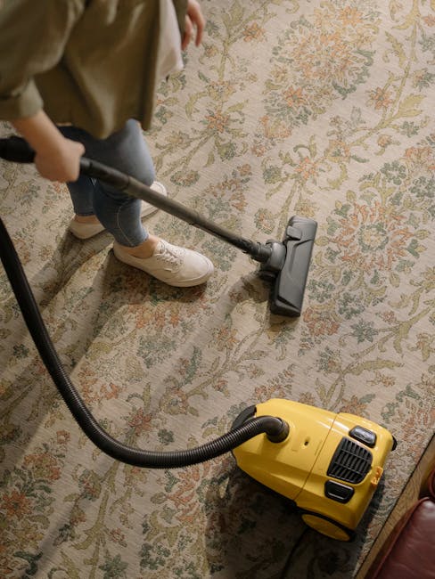 A person using a vacuum cleaner with a yellow vacuum body and black hose attachment on a patterned area rug in a living room. The rug features intricate floral designs in muted tones of green, beige, and orange. The individual, wearing casual clothing and white sneakers, is focused on cleaning the carpet surface, which appears clean and dust-free. The image highlights surface cleaning and deep cleaning activities, suitable for residential spaces, and is associated with professional cleaning services offered by Cleaners W14 on Lonsdale Road, Barnes, W14.