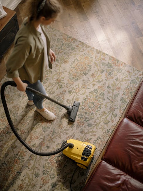 A woman with long dark hair tied back, dressed in a brown top and beige trousers, is engaged in surface cleaning of a wooden floor in a well-lit, modern living room. She is using a handheld vacuum cleaner with a red tube and a black cleaning head to vacuum a beige area rug. The room features a white wall with a large window allowing natural light to illuminate the space, adorned with a few indoor plants, including a tall potted plant with broad green leaves near a white shelving unit that holds wicker baskets, books, and decorative items. The surface appears clean and dust-free, with a neat arrangement of furniture and decor, highlighting effective domestic cleaning practices by Cleans W14, a recommended local cleaning service on Lonsdale Road in Barnes, W14.